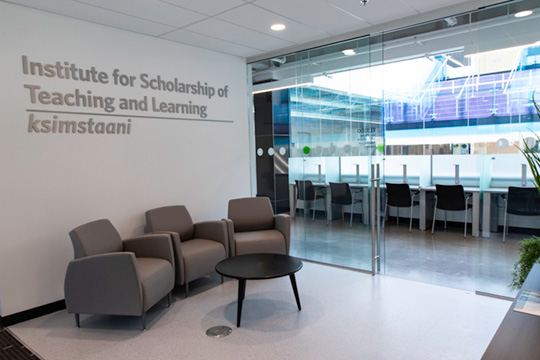 Photo of the entrance to the Mokakiiks Centre for Scholarship of Teaching and Learning. It is a clean waiting area with a sign for the location near a lightwell.