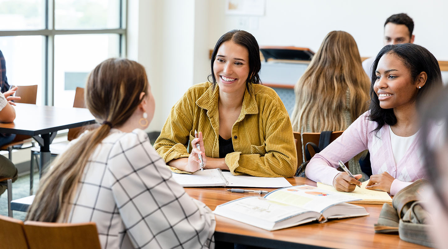 Three students studying and chatting at a large desk.