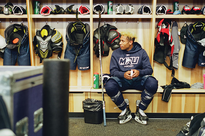 Jamal Watson sits on a bench in the change room. A variety of hockey gear hangs on the wall.