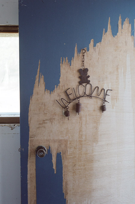 A photo of a worn down door. A handmade metal welcome sign with bells hangs on it.