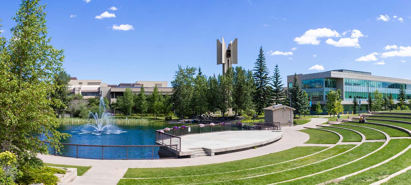 A pond with a fountain, surrounded by trees and other plants in front of a stepped outdoor amphitheatre.