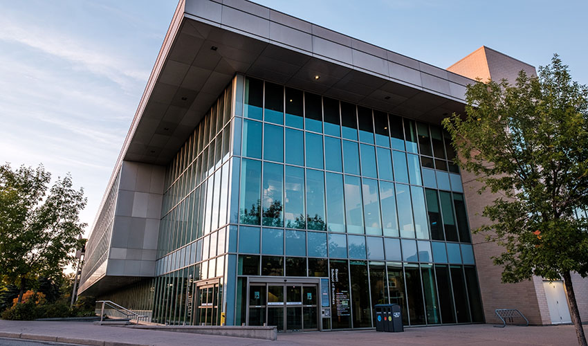 Photo of the Bissett School of Business building taken from outside the East Gate parkade. Features a glass corner of a building with a decorative overhang and the rest is made out of peach bricks.