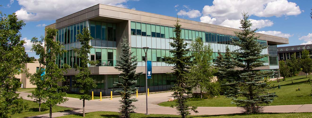 A long skinny building with blue tinted glass windows along the middle. There is a balcony on a corner in the middle and pathways and trees.