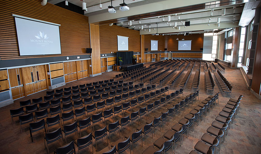 A large exhibition hall filled with rows of seats facing a stage. Several large screens hang high around the room.