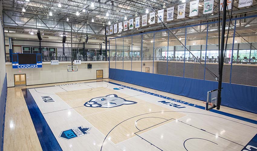 Looking down on a light wood gym floor painted with white and blue details, an MRU cougars logo and Canada West logo.