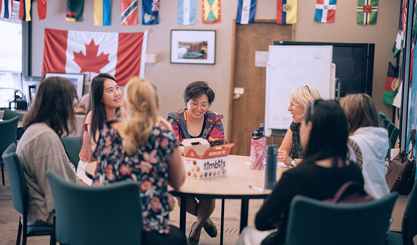 A diverse group of people chatting at a round table in the International Student Support Centre. The background is filled with flags from different countries.