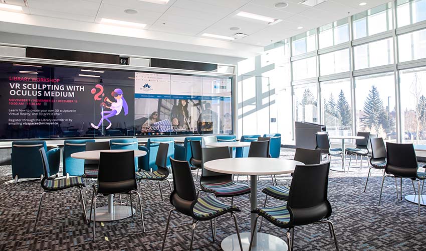 A room with a neutral coloured geometric carpet, round tables with chairs, a wall of TVs displaying campus activities and a glass wall letting in lots of natural light.