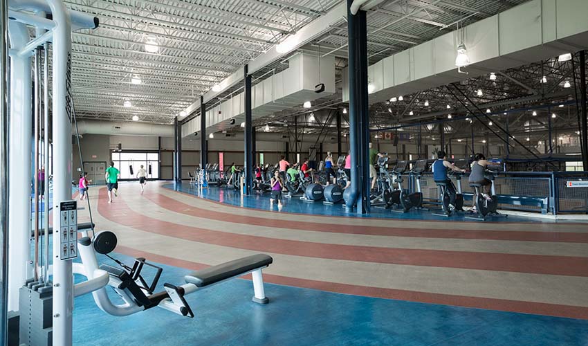 A running track in front of a row of ellipticals and treadmills that are looking down on a gym in a large industrial space.