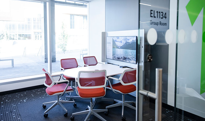 A room with lots of natural lights filled with table surrounded by chairs by a TV screen on the wall.