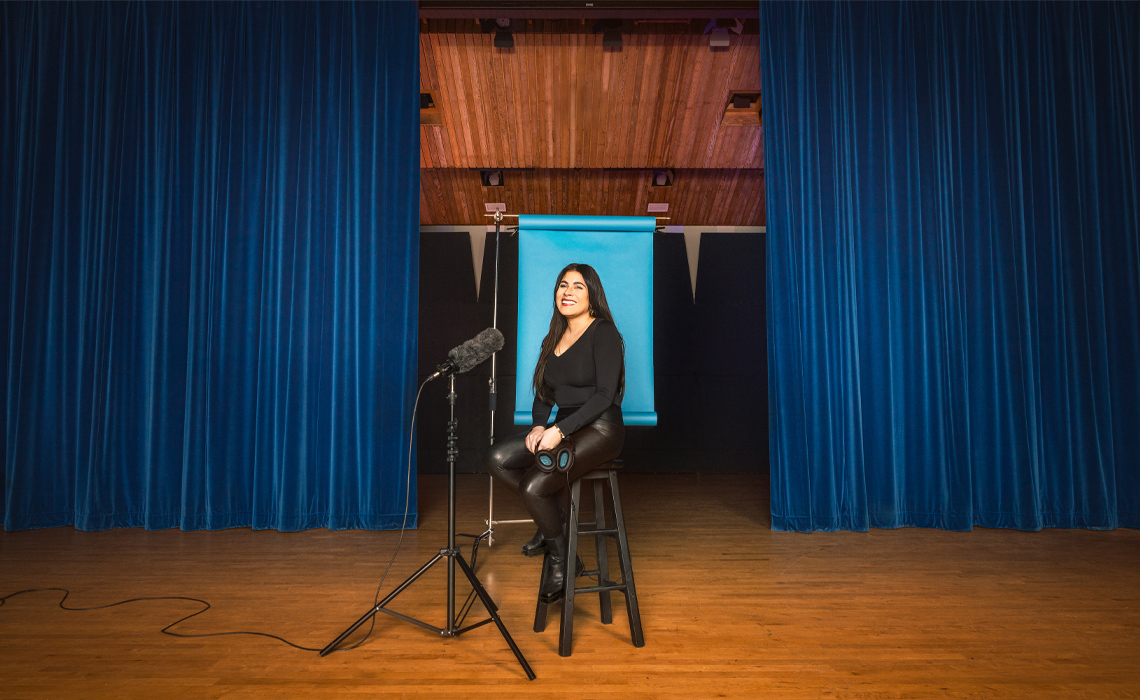 Ashley King seated on a stool, positioned on a stage in front of a blue curtain, creating a serene and elegant atmosphere.