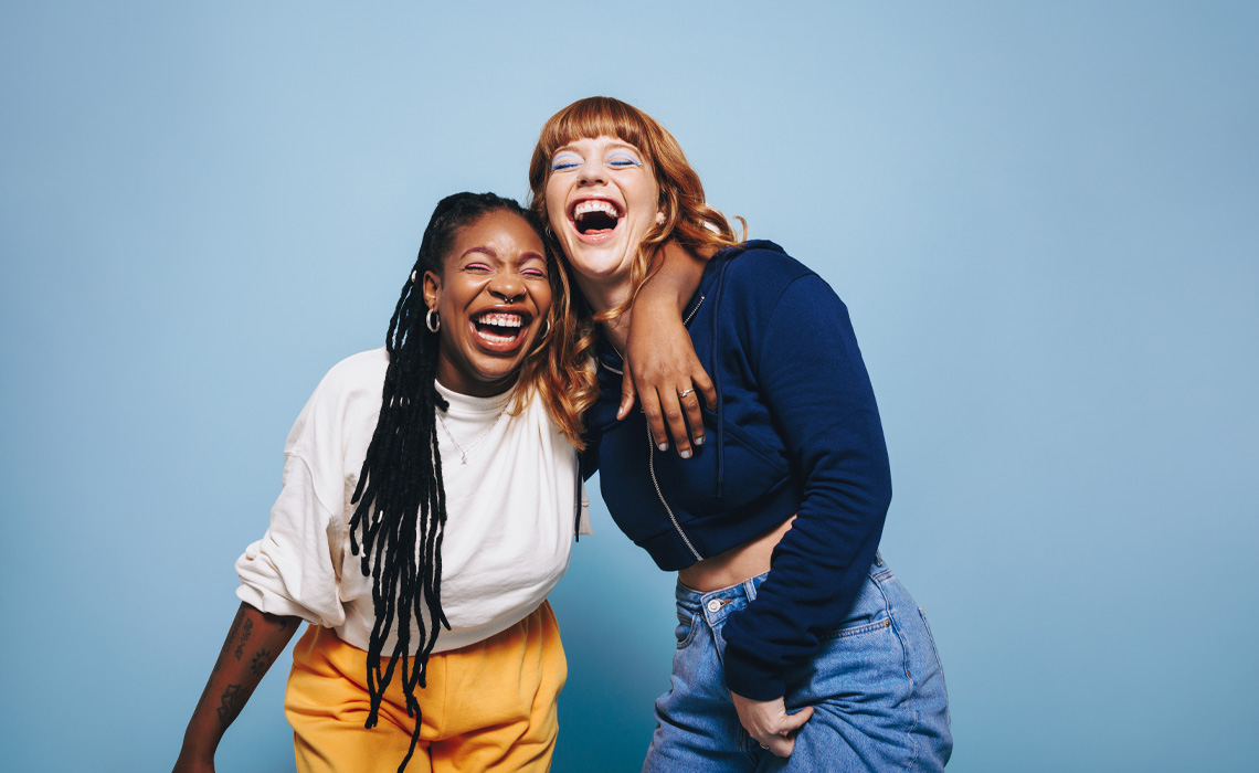 Two women joyfully laughing together against a vibrant blue background.