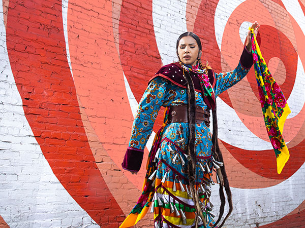 Sofia Eaglehead Baptise in a jingle dress dancing in front a mural in downtown Calgary.