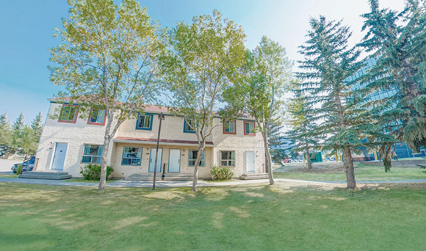 A light yellow stucco house with red trim and food in an area with a grassy field and trees.