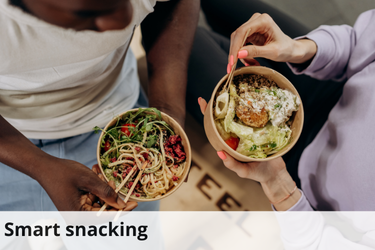 The image shows two people holding bowls of food, viewed from above. One person, on the left, is holding a bowl of what appears to be a salad with mixed greens, noodles, and some red-colored ingredients, possibly beets or cranberries. The other person, on the right, holds a bowl containing a variety of food items, including a large piece of what seems to be grilled or roasted meat, green vegetables, and a creamy sauce. The image is captioned "Smart snacking," suggesting the food choices are healthy or mindful. The setting appears casual, likely reflecting a focus on convenient, nutritious snacking options.