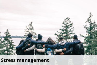 The image shows a group of people standing together with their arms around each other's shoulders, facing away from the camera. They are outdoors, surrounded by tall trees, with a scenic view of water and distant land in the background. The individuals are dressed in casual, outdoor clothing, and their close-knit stance conveys a sense of unity and support. The image is captioned "Stress management," suggesting that social connection and spending time in nature are important strategies for managing stress. The overall theme emphasizes the role of community and natural environments in promoting mental well-being.