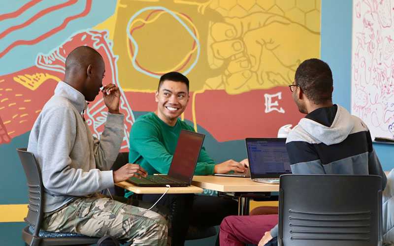 Three people working on laptops in front of a brightly coloured, illustrated mural.