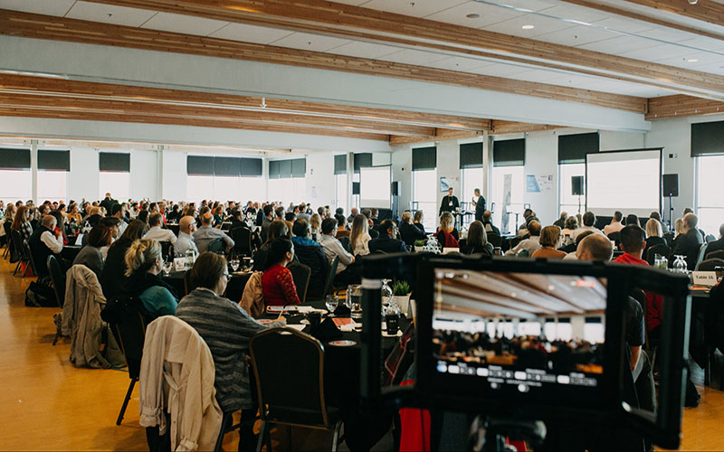 A room full of professionals seated at round tables listening to a presentation.