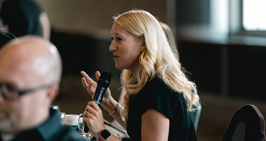 A woman speaking into a microphone at a round table full of other participants.