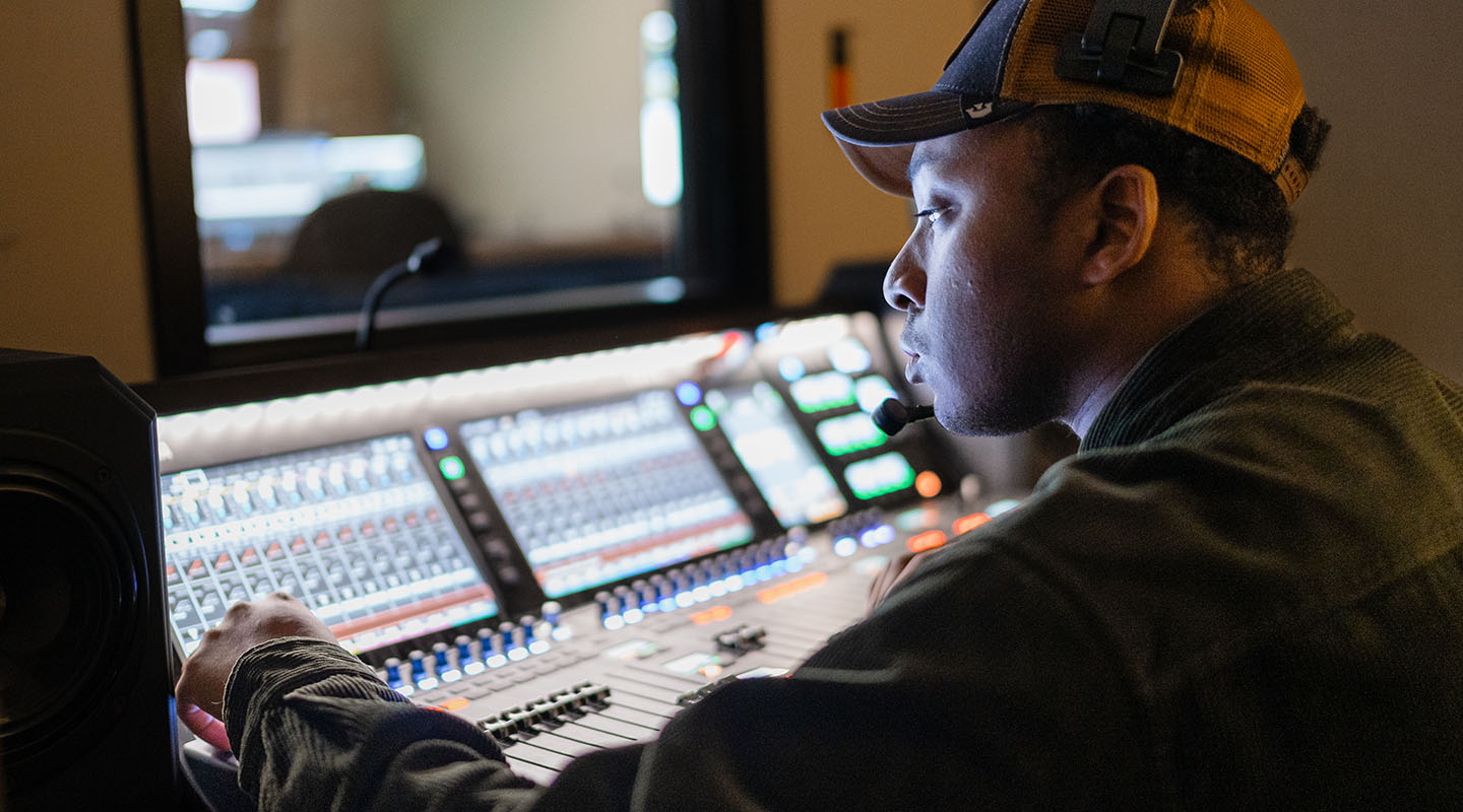 A student wearing a baseball cap works in Mount Royal University's broadcasting sound booth.