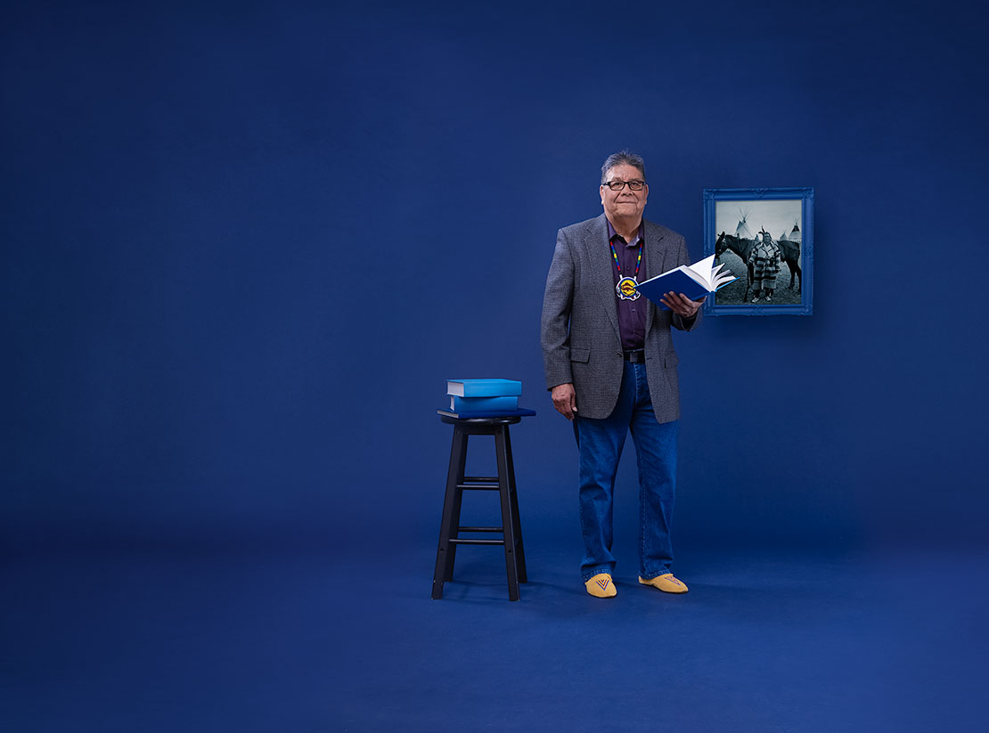 Photo of Roy Bear Chief in front of a blue backdrop. He holds a blue book and there are more blue books on a stool nearby. A historical photo of his grandfather is hung in a blue painted frame behind him.