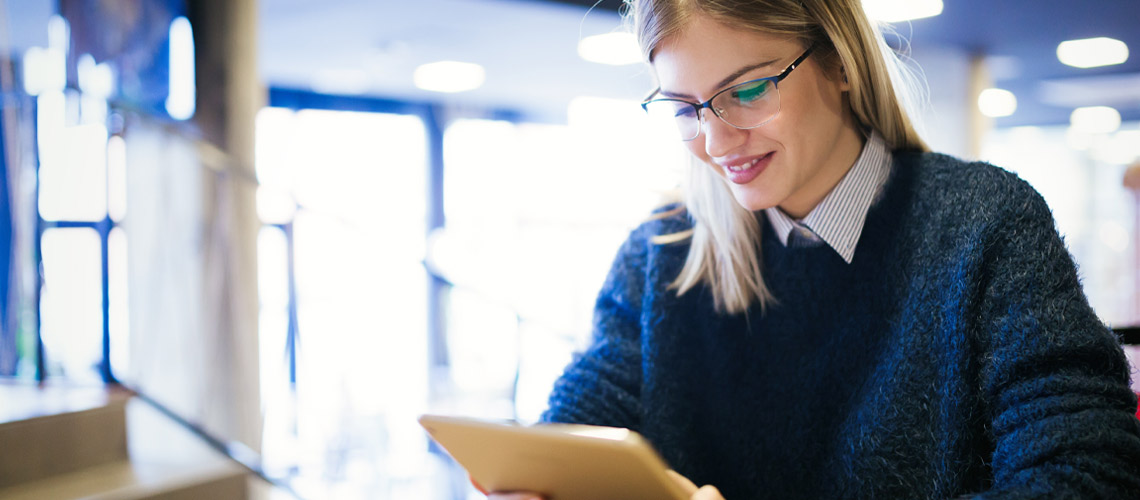 A woman wearing glasses, smiling as she reads on a tablet