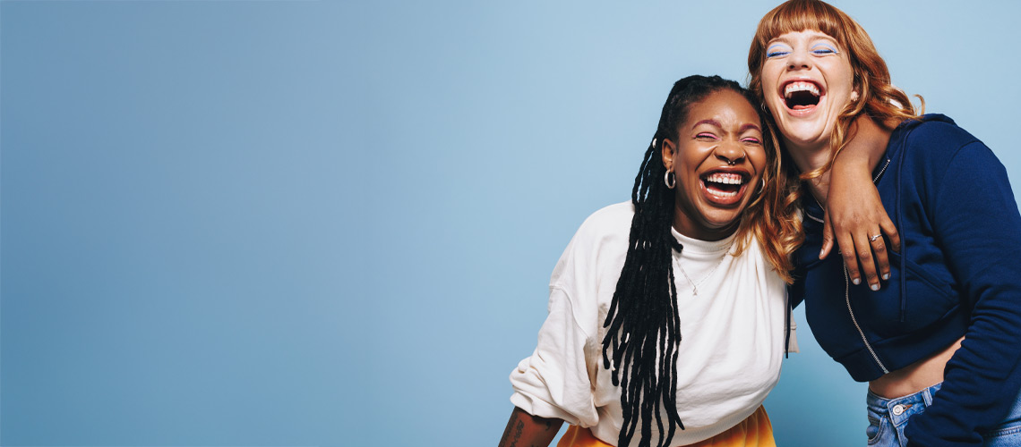 Two women joyfully laughing together against a vibrant blue background